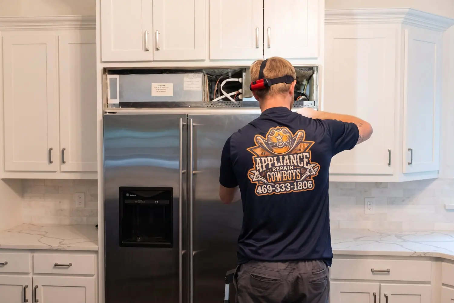Technician in a navy Appliance Repair Cowboys polo working on a built-in refrigerator in a white kitchen