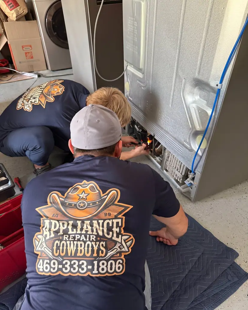 Two technicians brazing sealed-system lines at the back of a refrigerator